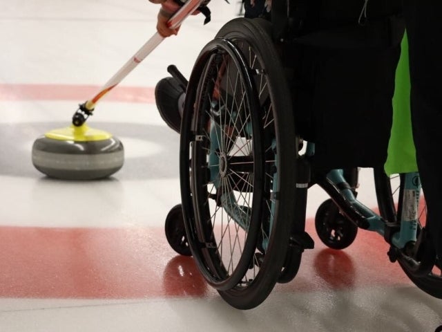 At a curling rink, we see a rock to the left and the back of a wheelchair with the person sitting in it taking their stick to move the rock