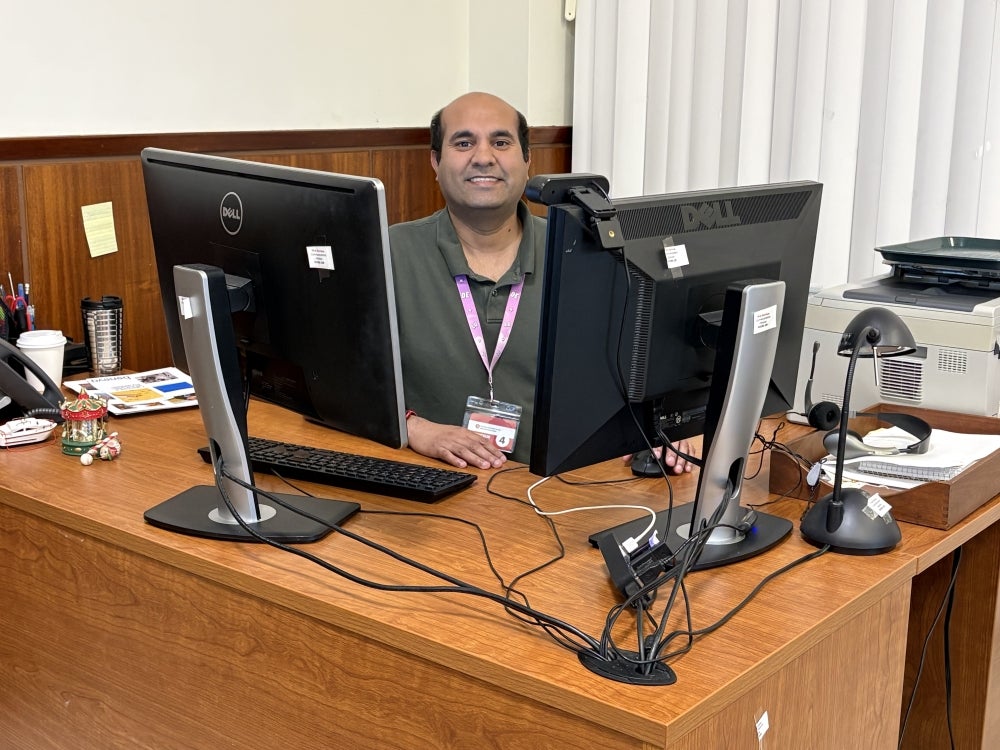 Fiaz Khalid sits between two computers, smiling and working, in te web office