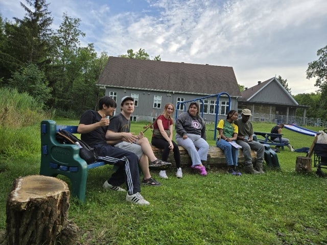 Several campers are seen seated, relaxing outside on a beautiful day at Camp de Grandpré in Otterburn Parkg a summer respite camp in 2025