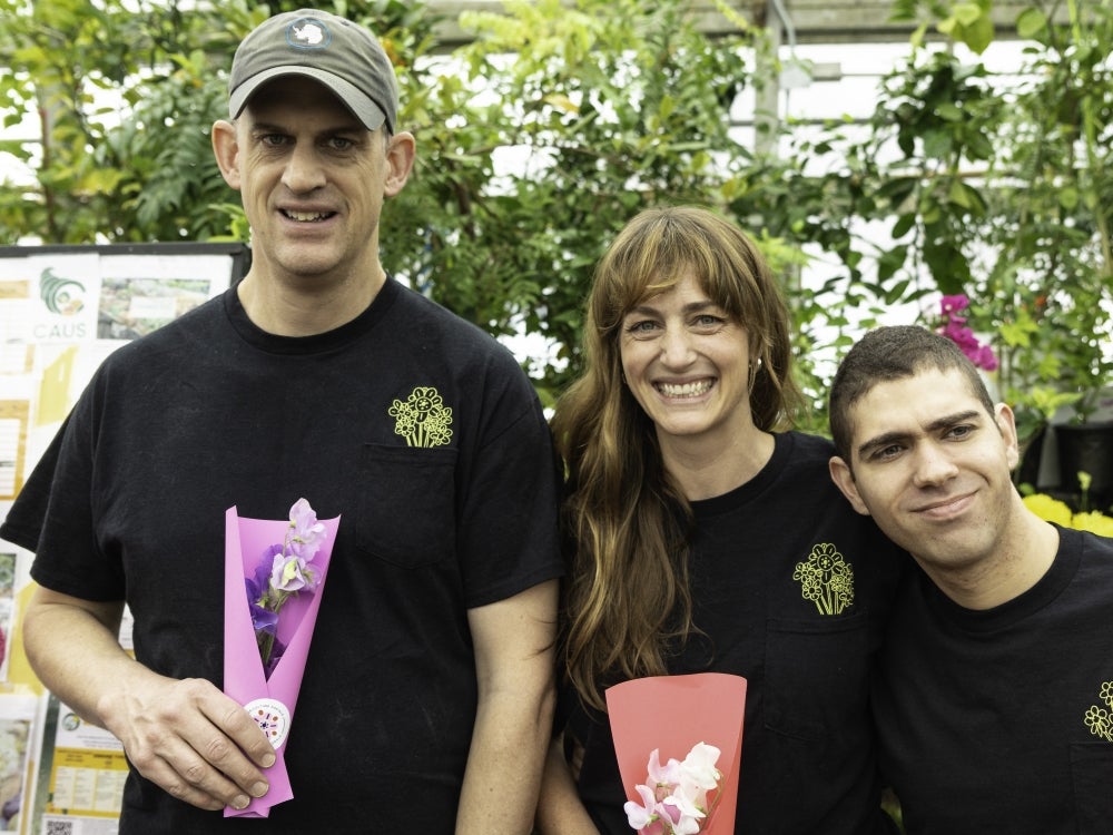 Harris, Lucy and Ellis stand facing the camera with green foliage in the background and holding colourful bouquets of flowers in their hands.