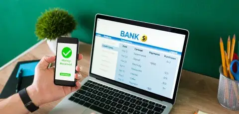 Persona caucasica revisando sus cuenta bancarias en la computadora y revisando su celular con la mano izquierda revisando su token digital.