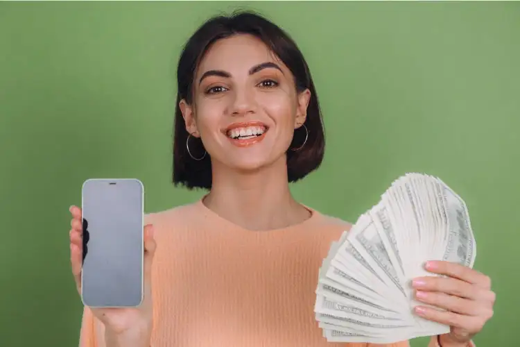 Mujer caucasica sonriendo y sosteniendo un celular en la mano derecha y billetes gracias a sus beneficios de tener una buena cuenta sueldo