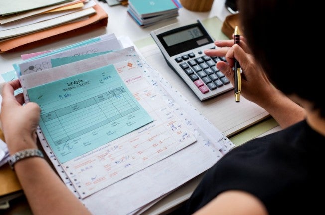 Mujer caucásica con una calculadora en la mano derecha y facturas en la mano izquierda calculando el igv correcto.