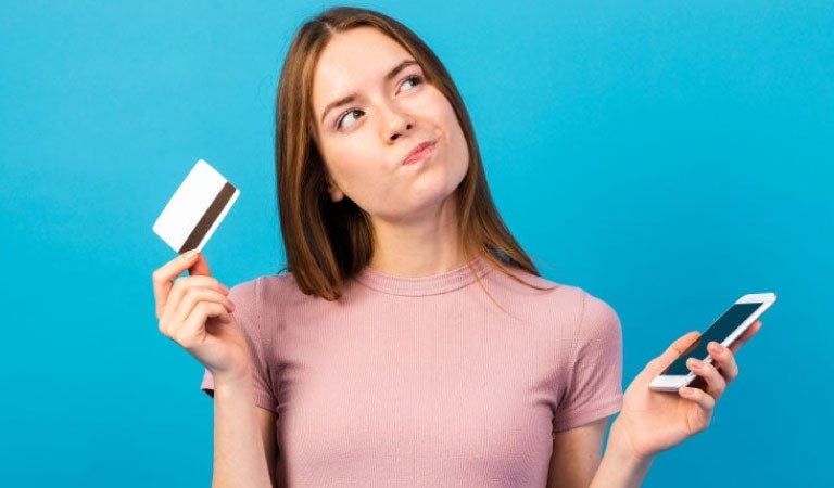 Mujer caucásica sosteniendo con la mano izquierda su teléfono celular y con la derecha una tarjeta de crédito pensando en que tarjeta debería usar, si la de crédito o la de débito