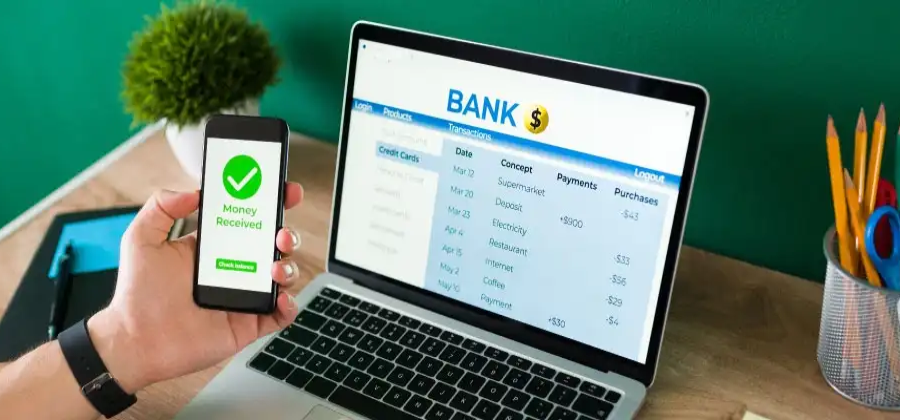 Persona caucasica revisando sus cuenta bancarias en la computadora y revisando su celular con la mano izquierda revisando su token digital.