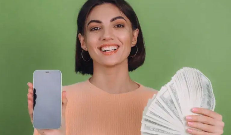 Mujer caucasica sonriendo y sosteniendo un celular en la mano derecha y billetes gracias a sus beneficios de tener una buena cuenta sueldo